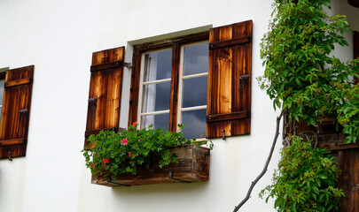 Fototapeta na wymiar a rustic window with cute white curtains, wooden shutters and red geraniums on the window ledge in the Bavarian alpine countryside Schwangau in the German Alps, Bavaria, Germany
