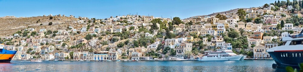 Fototapeta na wymiar Multi-colored houses scattered on hills of Symi island view from water. Coastal town on turquoise sea shore in Greece. Tourist liners moor by seafront