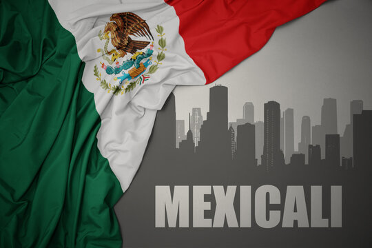 abstract silhouette of the city with text Mexicali near waving colorful national flag of mexico on a gray background.