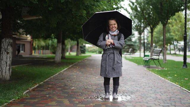 Joyful little person standing with umbrella on city alley looking up and looking at camera smiling stepping feet in rubber boots. Wide shot portrait of Caucasian little person posing on rainy day