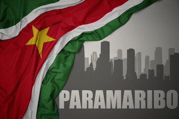 abstract silhouette of the city with text paramaribo near waving colorful national flag of suriname on a gray background.