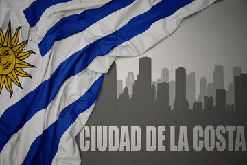 abstract silhouette of the city with text Ciudad de la Costa near waving colorful national flag of uruguay on a gray background.