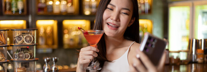 Young Asian woman takes a photo of herself or Selfie with a smiling face and drinks a cocktail in a vintage bar, Relaxing activities after work or hangouts, Place of entertainment for teens.