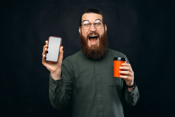 Amazed and shocked young man with beard showing smartphone screen and hodling cup of coffee take...