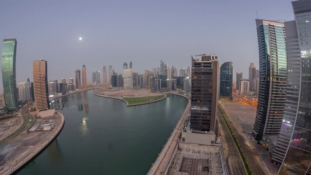 Cityscape panorama of skyscrapers in Dubai Business Bay with water canal aerial night to day transition timelapse with Moon setting down. Modern skyline with illuminated towers and waterfront before