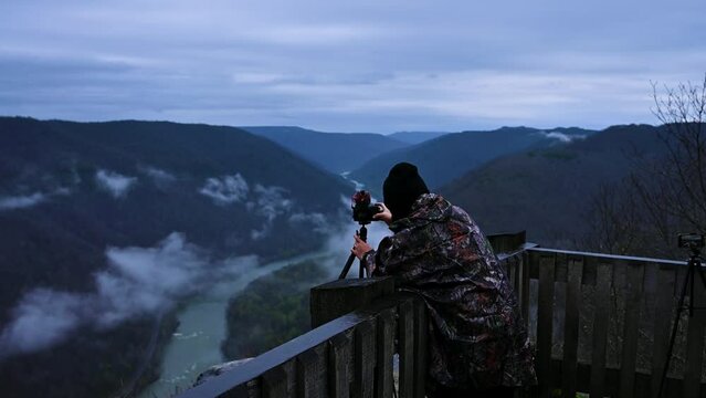 A boy is taking pictures of a mountain on a camera by the side of an iron bridge in a mountainous area