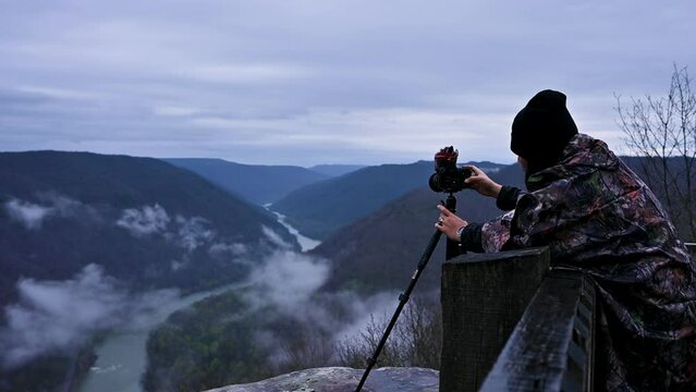A boy wearing a cap on his head and a jacket on his body is setting the camera on a tripod and taking pictures of the mountains with one mind