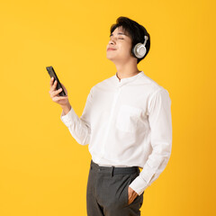 Young good looking asian man using smartphone and headphone listen to music isolated on yellow background