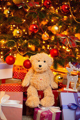 Cute teddy bear sitting on presents under Christmas tree. Beautiful composition of many boxes gifts for Christmas and a bear toy among them beneath Christmas tree