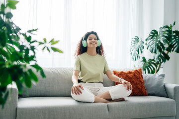 Relaxed young calm woman sitting at home at the sofa in lotus position, while meditating and...