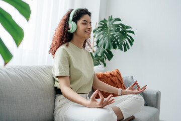 Happy young woman meditating at home in the morning and practicing yoga while sitting at the sofa. Woman breathing and listening music while sitting indoor in lotus pose