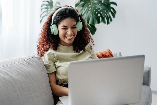 Attractive Young Woman Sitting On The Sofa And Using Her Laptop And Headphones To Make A Video Chat With Someone At Home. Multiracial Girl Laughing Out Loud