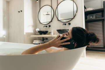 Brunette pretty woman speaking by phone while laying and relaxing at the bathroom indoor. Body care and self care routine concept