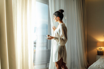 Young woman wearing white bathrobe opening curtains in luxury hotel room and enjoying sun in the morning while having her cup of coffee - obrazy, fototapety, plakaty