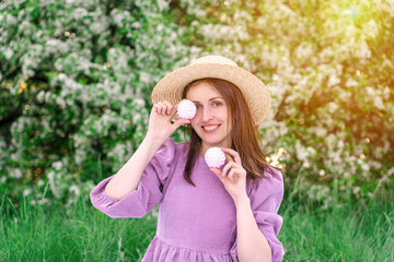 A pretty young woman in a straw hat is fooling around with marshmallows at a picnic in a blooming apple orchard