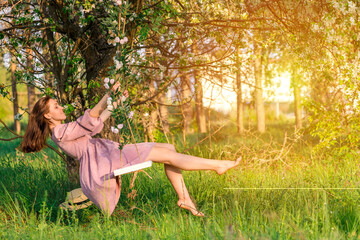 A brunette woman in a purple dress swings on a rope swing in an apple orchard at sunset