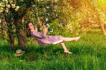 A brunette woman in a purple dress poses on a rope swing in an apple orchard at sunset
