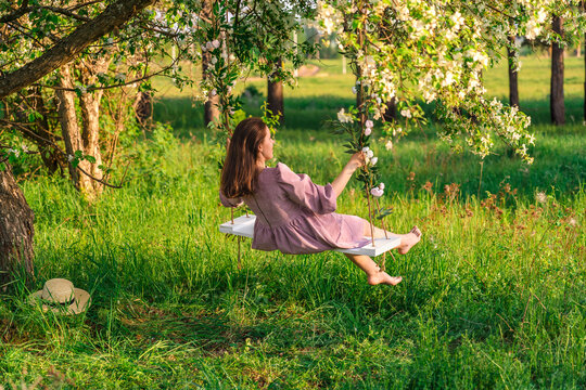 A brunette woman in a purple dress enjoys swinging on a rope swing in an apple orchard at sunset