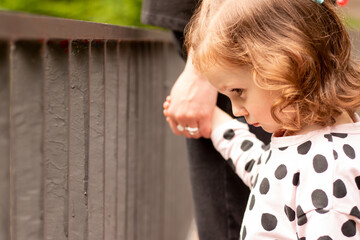 Little beautiful red-haired girl with curly hair in a dress holds the hand of a parent in a summer park