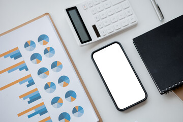 An image of the top corner of a desk with financial documents showing charts, a white calculator, a notebook and pen, a blank mobile phone screen for advertising messages. copy space.