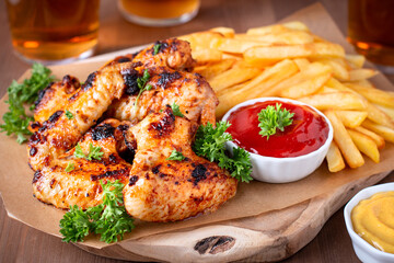 Roasted chicken wings and fries on wooden board and beer in background