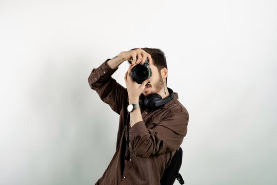 Young man wearing brown shirt posing isolated over white background taking images with dslr camera. Photographer covering his face with the camera.