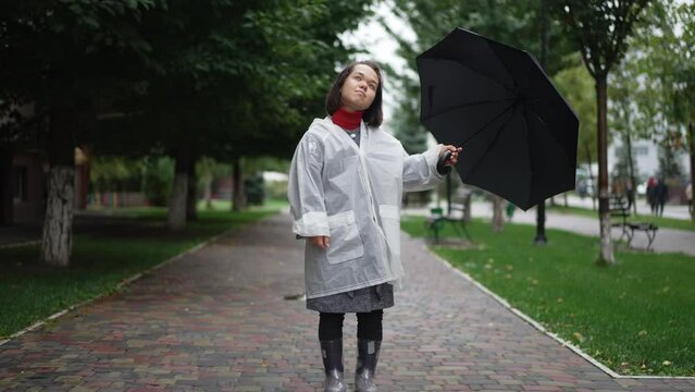 Wide shot young beautiful little woman in rain coat and rubber boots thinking looking up standing with umbrella outdoors. Portrait of Caucasian little person dreaming resting on city street