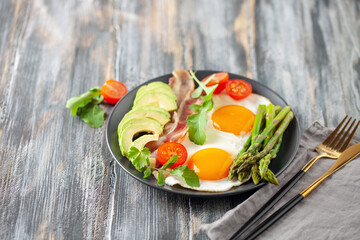Tasty breakfast. Fried eggs with green asparagus, fried bacon on black plate on wooden table.