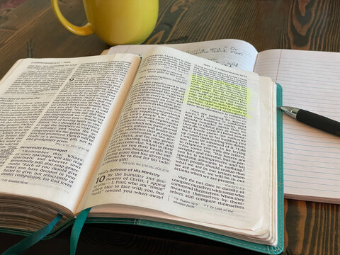 Open Bible laying on table with highlighted passage. II Corinthians 10. Bible laying open with teal colored book marks. Passage highlighted in yellow. Yellow coffee mug, notebook and pen in background