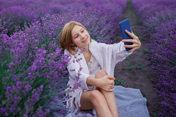 Teenage girl enjoys lavender field and make photo with smartphone. Gen Z teenager with gadget walks among purple flowers at sunset. Technology and beauty of nature, summer travel and vacation