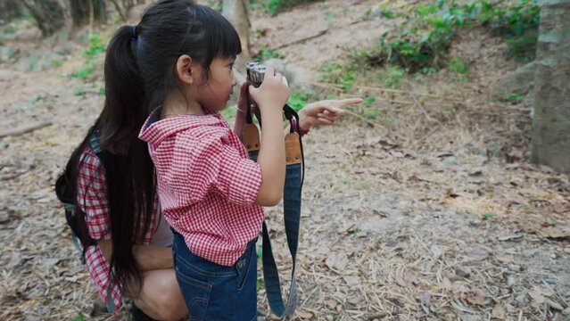 Side view of Little cute Asian girl with bangs in red plaid shirt and denim bib standing next to Asian mother among forest trees, raise the vintage film camera taking a photo of nature in front of her