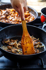 Fried champignon mushrooms with onions in a frying pan with a wooden spatula stirs a female hand.