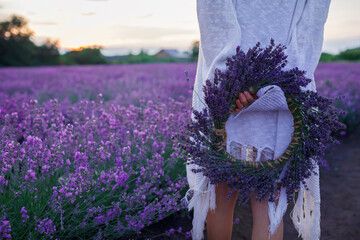 Young girl holds wreath of lavender flowers behind her back and enjoys beautiful sunset over the field. Happy teenager among purple flowers. Calm landscape, beauty of nature, summer travel, vacation