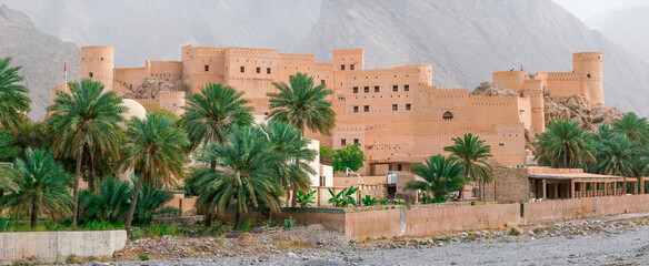 Nakhal, Oman - 04.02.2018: Panoramic shot of medieval Arabian military fortress. Very hot day with heat haze in the sky. Old Arabian castle. Tall stone walls. Palm trees in the front.