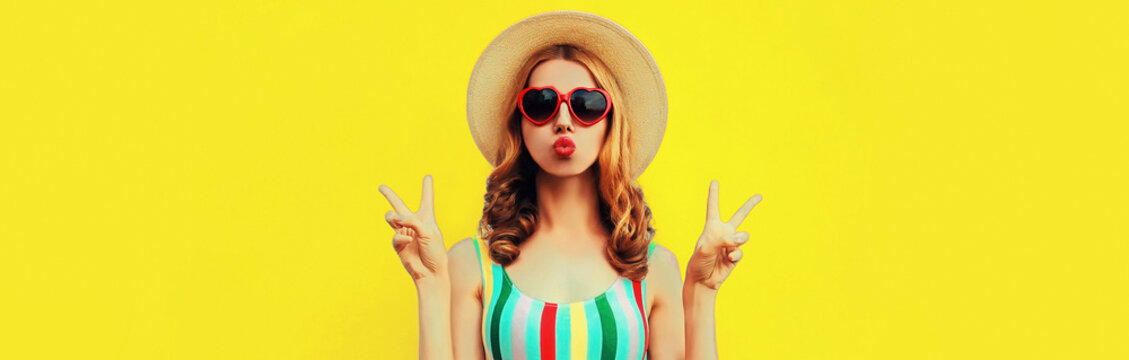 Portrait of beautiful young woman model blowing her lips and posing wearing summer straw hat, red heart shaped sunglasses on yellow background, blank copy space for advertising text