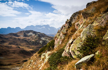 Rocks, bushes and dry grass are seen with rocky mountain peaks in the background during sunny weather in national park in Caucasus nature reserve. Alpine landscape