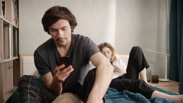A couple sits in bed with a phone in their hands, then the guy shows his phone and they smile. Slow motion 4k footage