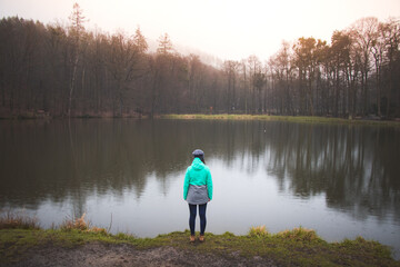 Fototapeta na wymiar Hiker in a grey and blue jacket at the edge of a pond in rainy weather, enjoying the misty sunset light. Biodiversity of Czech nature. Young pretty brunette discovering new landscapes