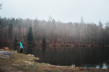 Fototapeta na wymiar Hiker in a grey and blue jacket at the edge of a pond in rainy weather, enjoying the misty sunset light. Biodiversity of Czech nature. Young pretty brunette discovering new landscapes