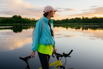 Woman is standing with mountain bike in cross country road at sunset in summer. Looking direct. Colorful lake landscape with sporty girl with backpack riding bicycle. Sport and travel. - 512433404
