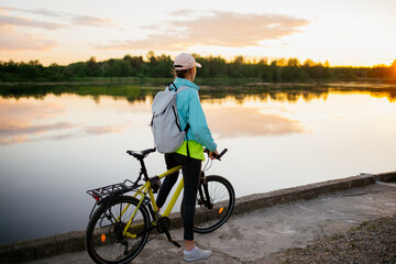 Woman is standing with mountain bike in cross country road at sunset in summer. Looking direct. Colorful lake landscape with sporty girl with backpack riding bicycle. Sport and travel. - 512433445