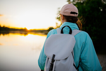 Traveling woman with backpack and hat looking at river at sunset. View from front tourist traveler bag. - 512433477