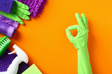 Female hand shows OK gesture over orange background with cleaning supplies and chemicals.