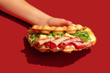 Hand holding bubble waffle sandwich with ham, tomato, lettuce, bacon, sauce, mustard, cheese, red...