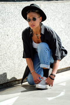 Photo of stylish young girl in fashionable sunglasses, jeans and black hat ties shoe laces on the city street