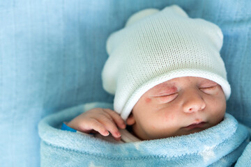 Newborn Baby Red Cute Face Portrait Early Days Sleeping In Medical Glass Bed On Blue Background. Child At Start Minutes Of Life On Hat. Infants, Childbirth, First Moments Of Borning, Beginning Concept