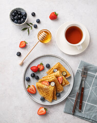 Homemade thin pancakes or crepes with fresh blueberries and strawberries, honey and cup of tea on a light background. Top view