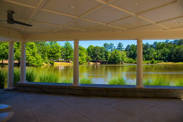 a white pergola with round pillars on the banks of a silky green pond with ceiling fans and round...