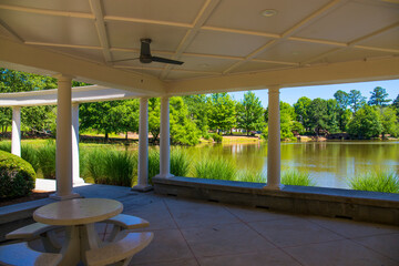 a white pergola with round pillars on the banks of a silky green pond with ceiling fans and round...