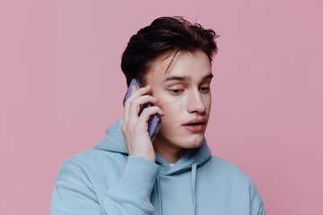 a handsome, thoughtful man is standing on a pink background in a light blue hoodie and holding a smartphone in his hands talking on it. Horizontal Studio Photography